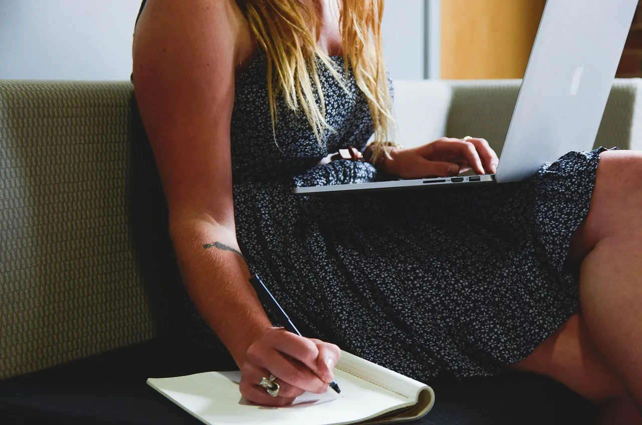 Frau in Sommerkleid schreibt Notizen, Laptop auf dem Schoß.