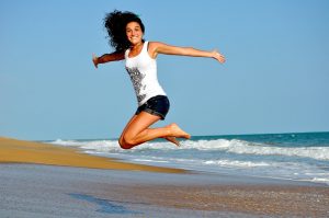 Auf dem Bild sieht man eine Frau am Strand die gerade in die Luftsprünge und sehr lächelt.