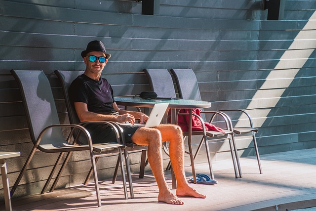 Mann sitzt mit Sonnenhut, Brille und Laptop am Pool