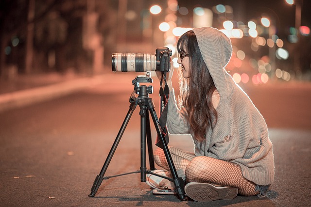 Brünette junge Frau mit grauem Hoodie und Kapuze über dem Kopf und Sonnenbrille sitzt nachts Lächelnd mit Fotostativ auf Straße.