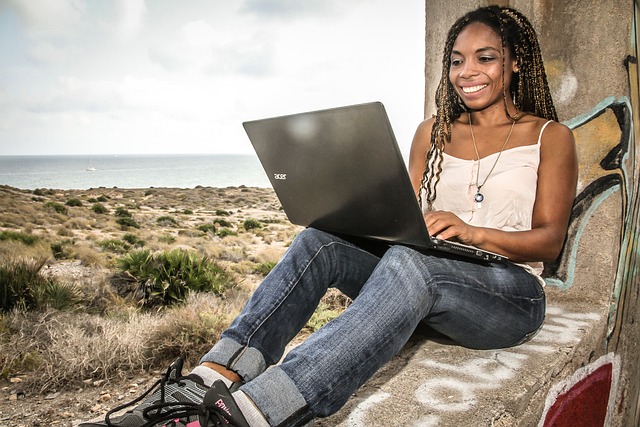 Frau sitzt lächelnd an Steinwand gelehnt mit Laptop, im Hintergrund Steppenlandschaft und das Meer.