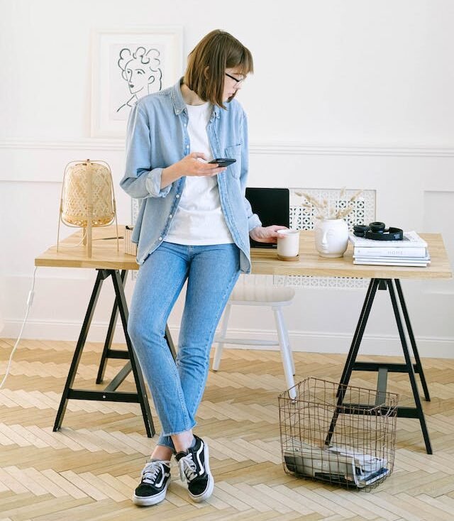 Dunkelblonde Frau mit Bob-Frisur steht vor Laptop-Tisch mit Handy in der Hand. Hintergrund ist weiß.