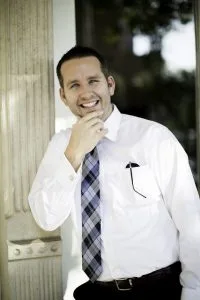 Mann im weißen Anzug und karierter Krawatte lächelt glücklich und hält sich am Kinn. Im Hintergrund eine antike Säule und Bäume.