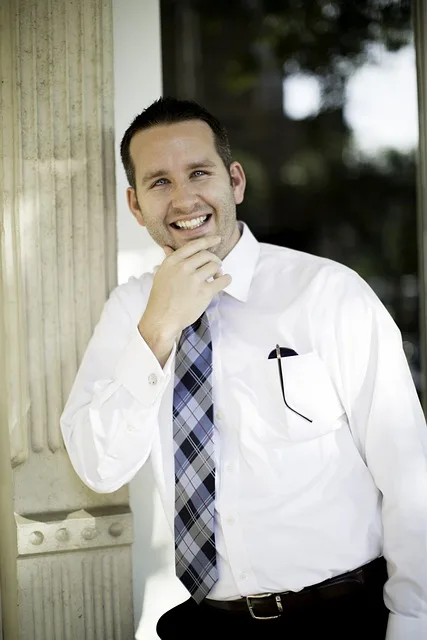 Mann im weißen Anzug und karierter Krawatte lächelt glücklich und hält sich am Kinn. Im Hintergrund eine antike Säule und Bäume.