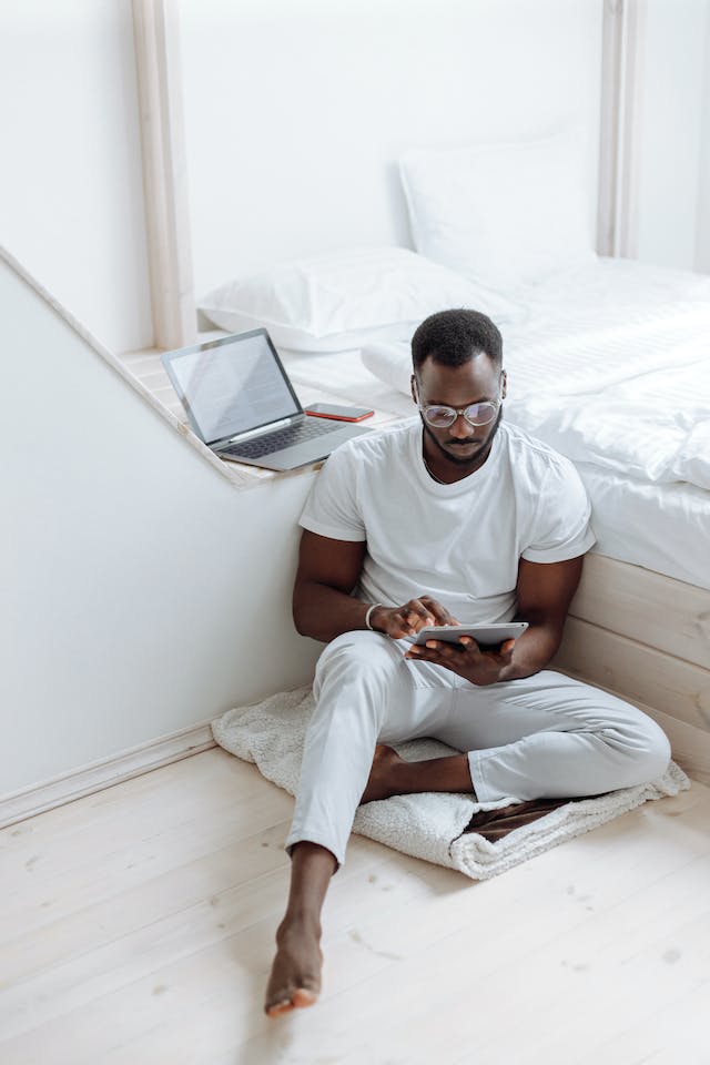 Mann mit kurzem schwarzem Haar, Drei-Tage-Bart und Brille sitzt vor einem weißen Bett und hält ein Tablet in der Hand. Hinter ihm ist ein Laptop.