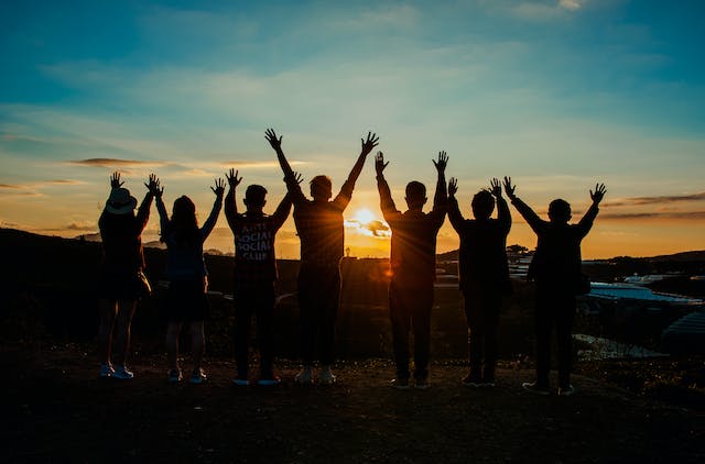 Silhouetten von Menschen mit feierlich erhobenen Händen vor einem Sonnenuntergang.