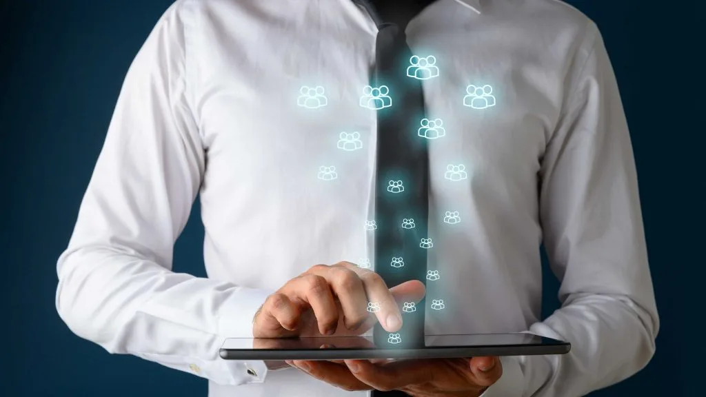 Vorderansicht eines Geschäftsmannes mit weißem Hemd und dunkler Krawatte, der ein digitales Tablet verwendet, aus dem viele leuchtende Menschensymbole herauskommen.