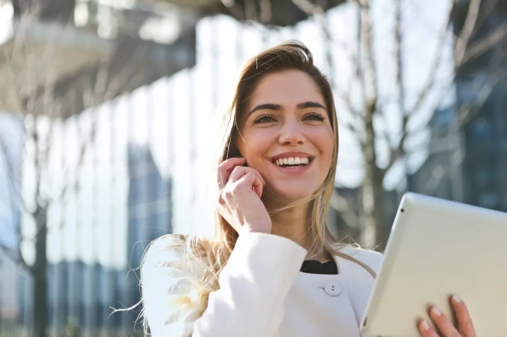 Blonde Frau mit weißem Blazer hält ein Tablet in der einen und telefoniert mit Handy in der anderen Hand. Sie ist mitten in der Stadt. Im Hintergrund Bäume und ein verschwommenes Gebäude.