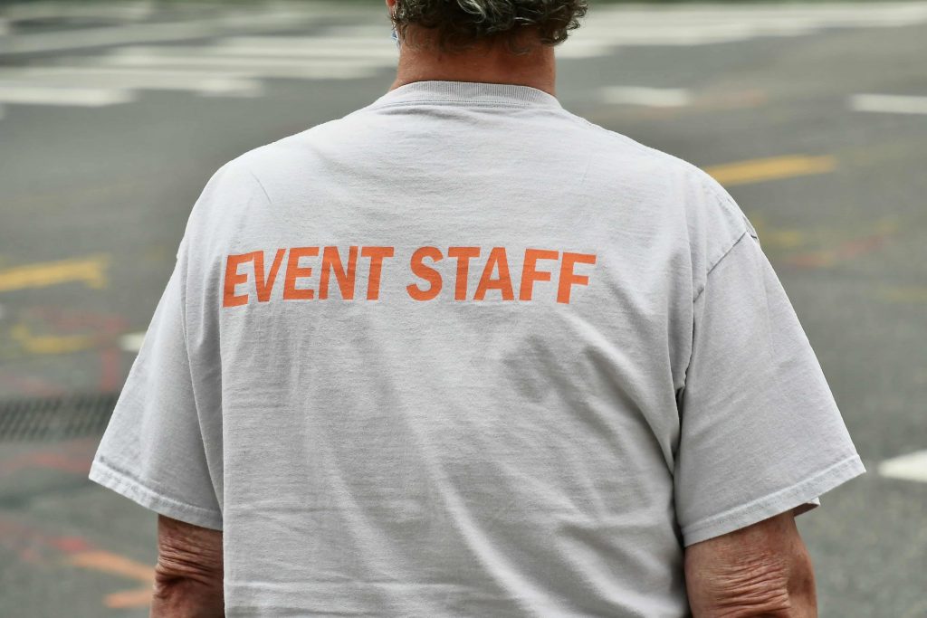 Ein männlicher Event-Mitarbeiter mit dunklen Locken mit dem Rücken zum Bild, trägt ein weißes Shirt, auf dem in oranger Schrift „Event Staff“ geschrieben steht.