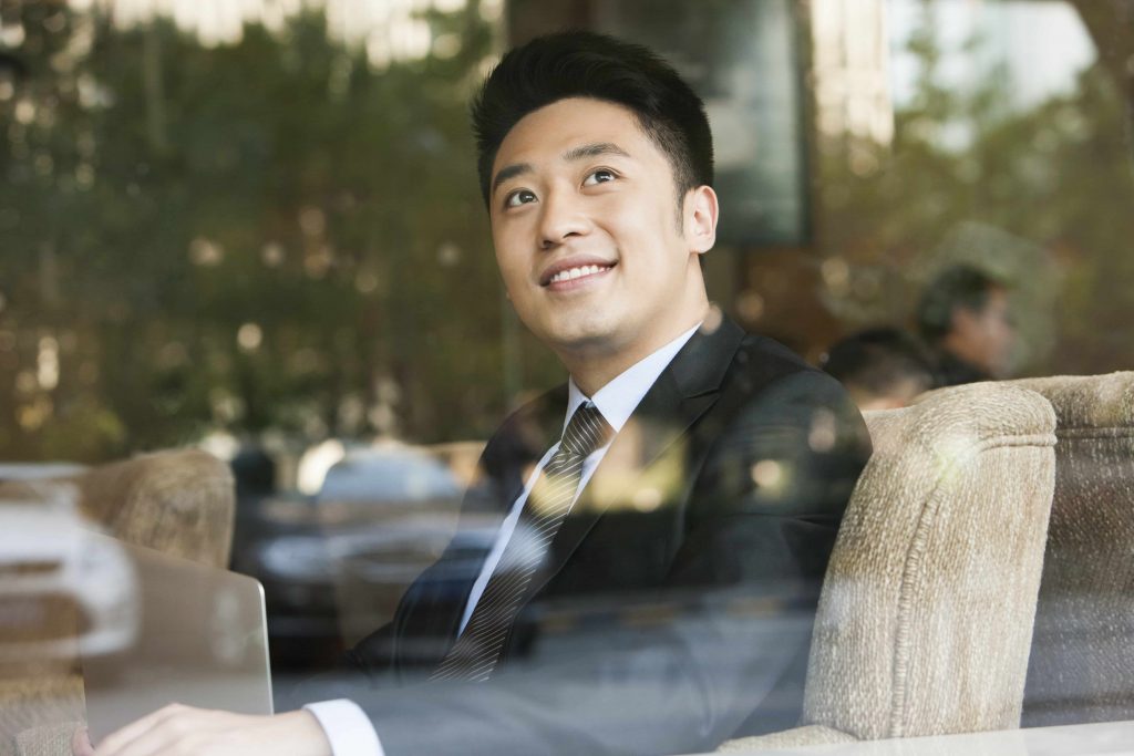 Mann in Business-Anzug auf einem braunen Sessel blickt lächelnd aus einem Fenster, an dem sich Laubbäume widerspiegeln.