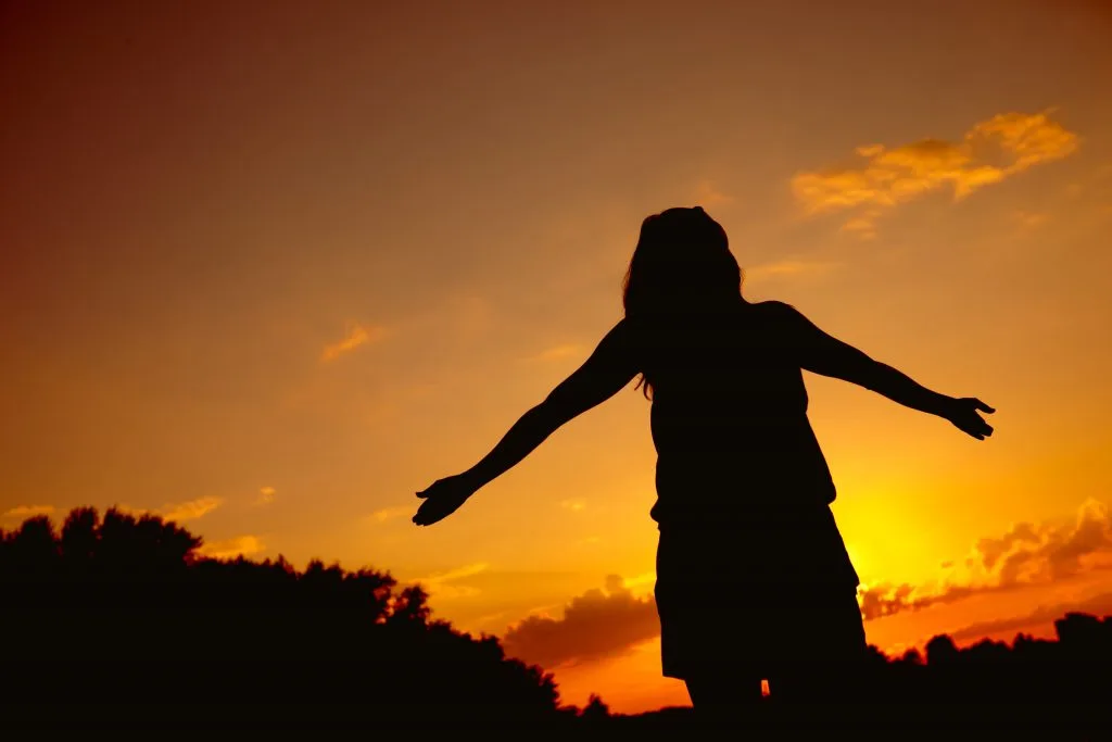 Silhouette einer Frau mit langem Haar, die mit ausgebreiteten Armen unter einer tieforangen Abenddämmerung steht. Im Hintergrund Bäume.