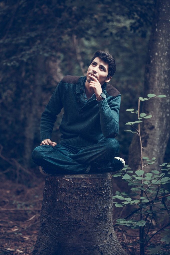 Dunkelhaariger Mann in dunkler Kleidung sitzt im Schneidersitz auf Baumstamm in Wald und hält nachdenklich den Finger ans Kinn.