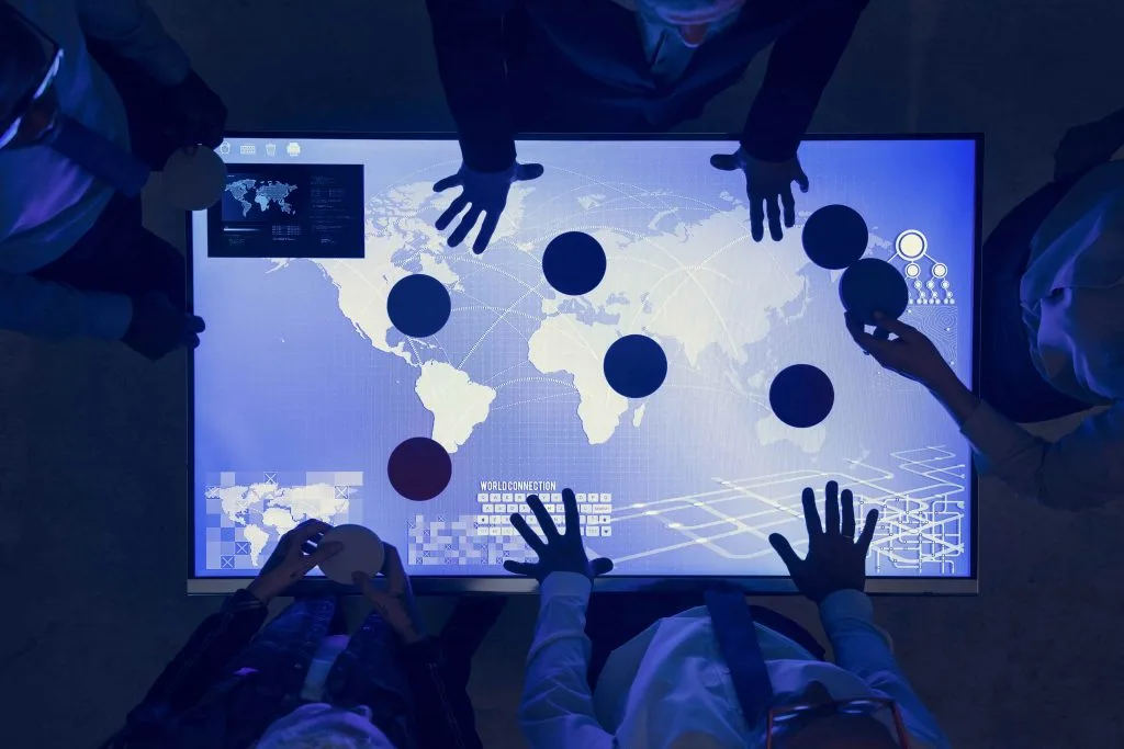 Mehrere Handpaare fassen an einen leuchtend-blauen Bildschirm, der eine vernetzte Weltkarte mit Punkten auf einigen Ländern zeigt.