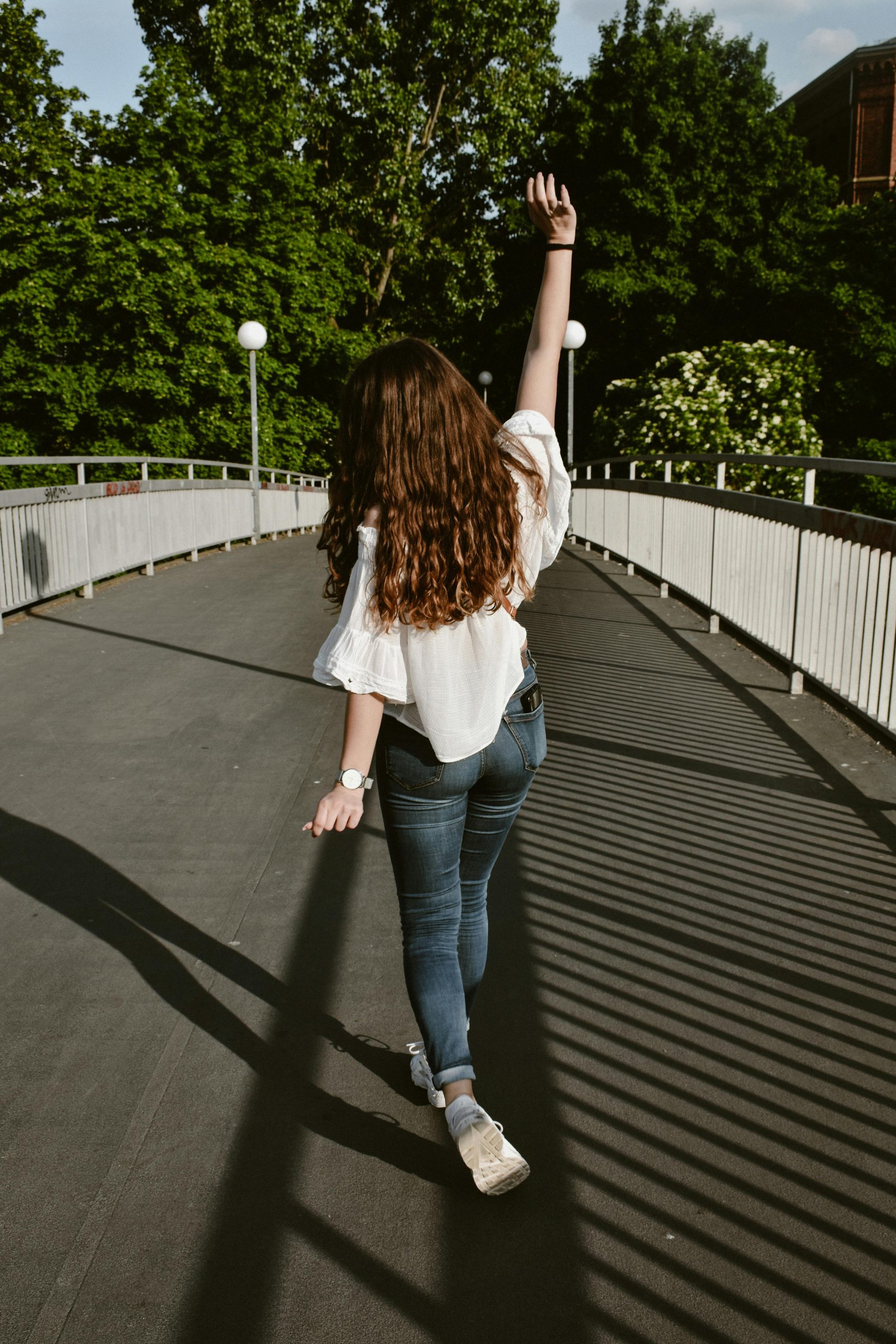 Brünette Frau von hinten in weißer Bluse und Jeans stemmt eine Hand hoch und läuft über eine Brücke auf Bäume zu. Strahlend blauer Himmel darüber.