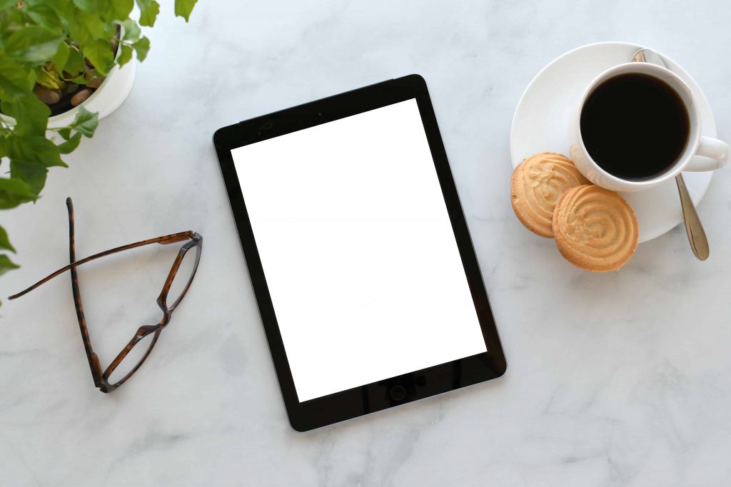 E-Reader mit weiß-leuchtendem Bildschirm liegt auf weißer Marmortischplatte, daneben eine Brille und eine Kaffeetasse mit zwei Keksen. Dahinter eine Zimmerpflanze.