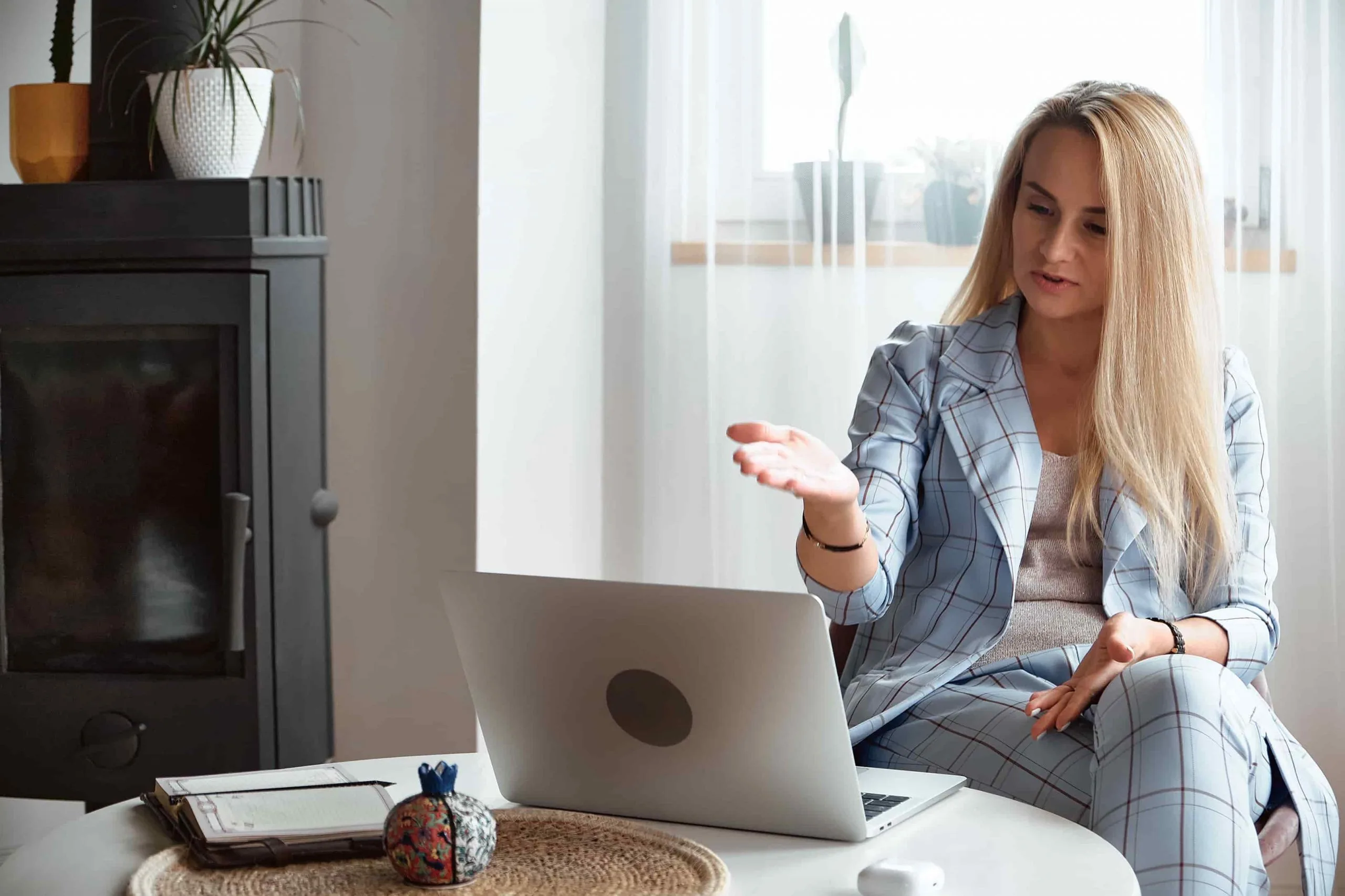 Blonde Frau mit blau-kariertem Kostüm sitzt vor einem runden weißen Tisch und erklärt etwas während einer Konferenz am Laptop. Im Hintergrund hellbraune Wand, Fenster mit weißen Gardinen, schwarzer Kamin und Zimmerpflanzen.