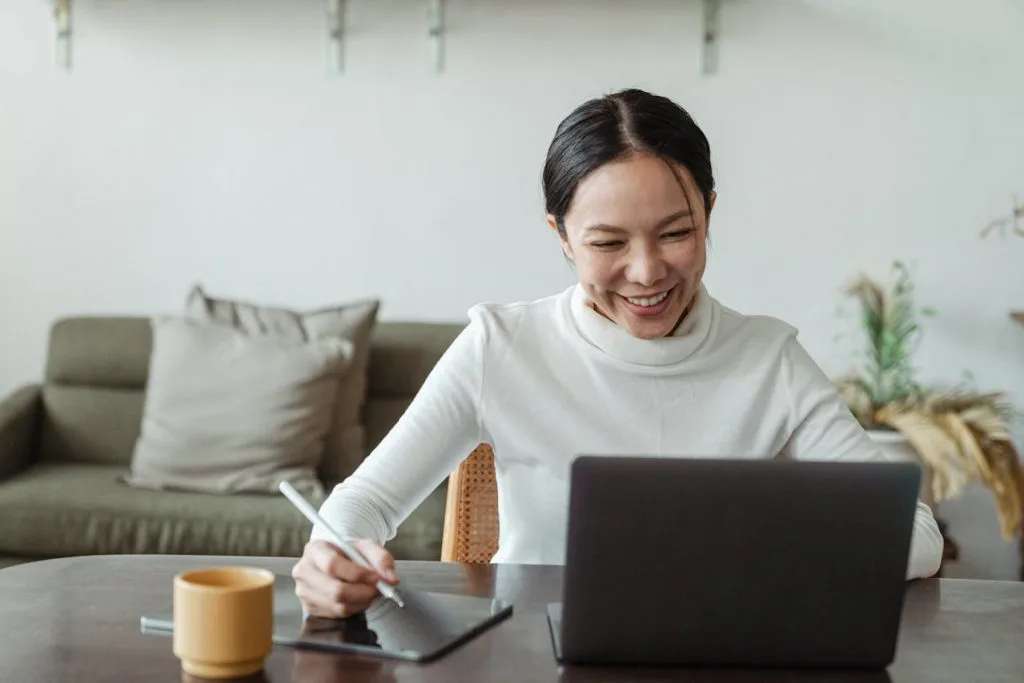 Frau mit dunklem zusammengebundenem Haar und weißem Pullover sitzt am Schreibtisch mit Laptop und Kaffee, lächelt und macht sich nebenbei Notizen. Im Hintergrund grüne Couch, Zimmerpflanze und weiße Wand.