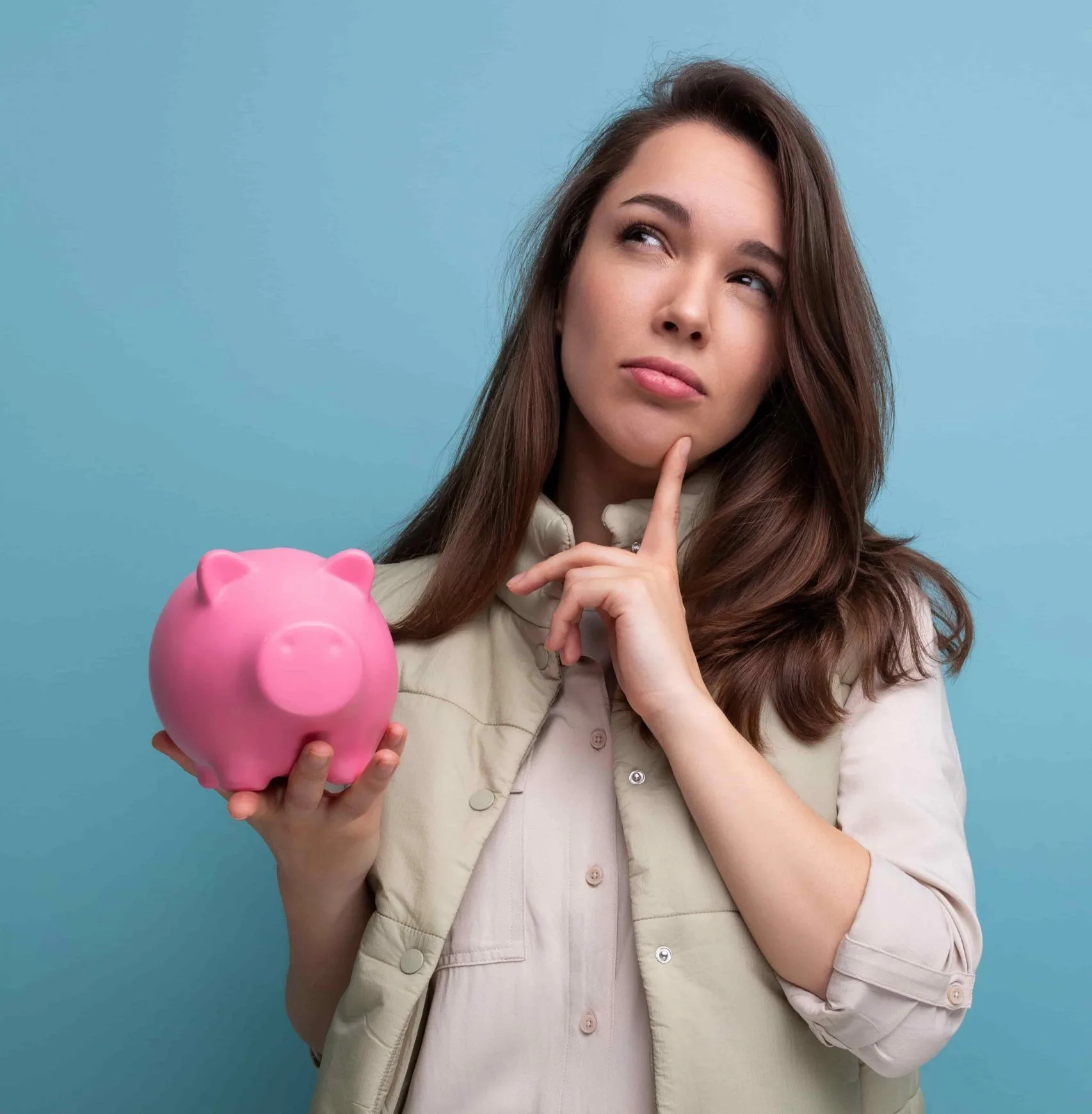 Brünette Frau in heller Bluse und Weste hält ein rosa Sparschwein in der Hand und überlegt. Blauer Hintergrund.