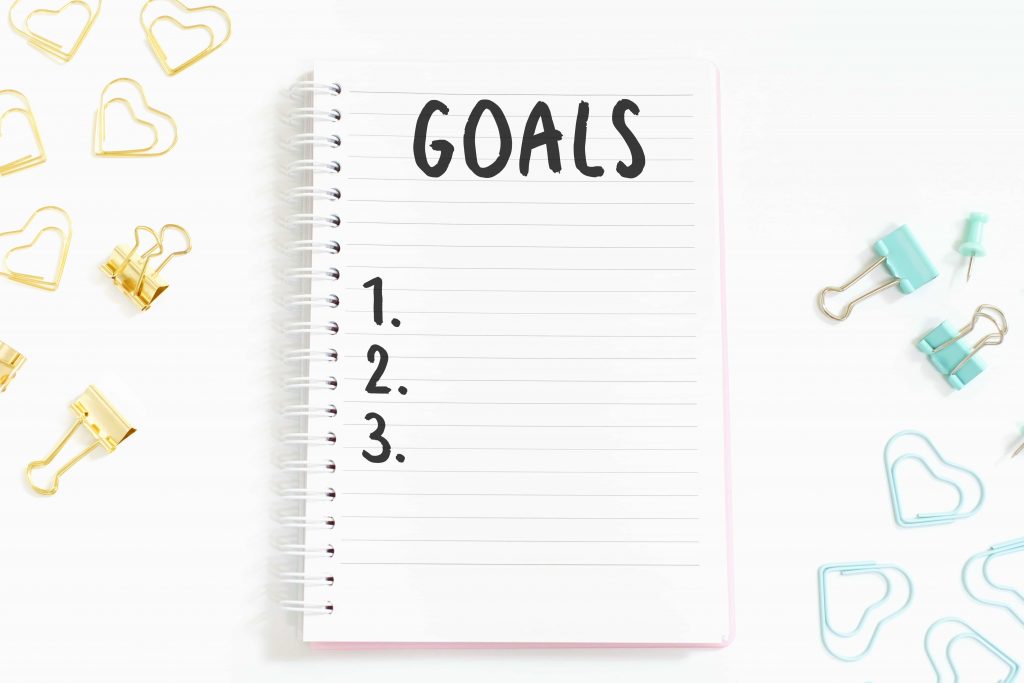 Das Wort Goal und Nummern darunter auf Notizblock auf weißem Hintergrund, Ansicht von oben. Daneben goldene und Blaue Heftklammern. 