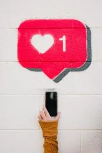 Ein roter Like-Button mit Herz auf einer weißen Wand gesprayt. Davor ein Arm mit Ring am Finger und orangen Ärmel, der ein Smartphone hochhält.