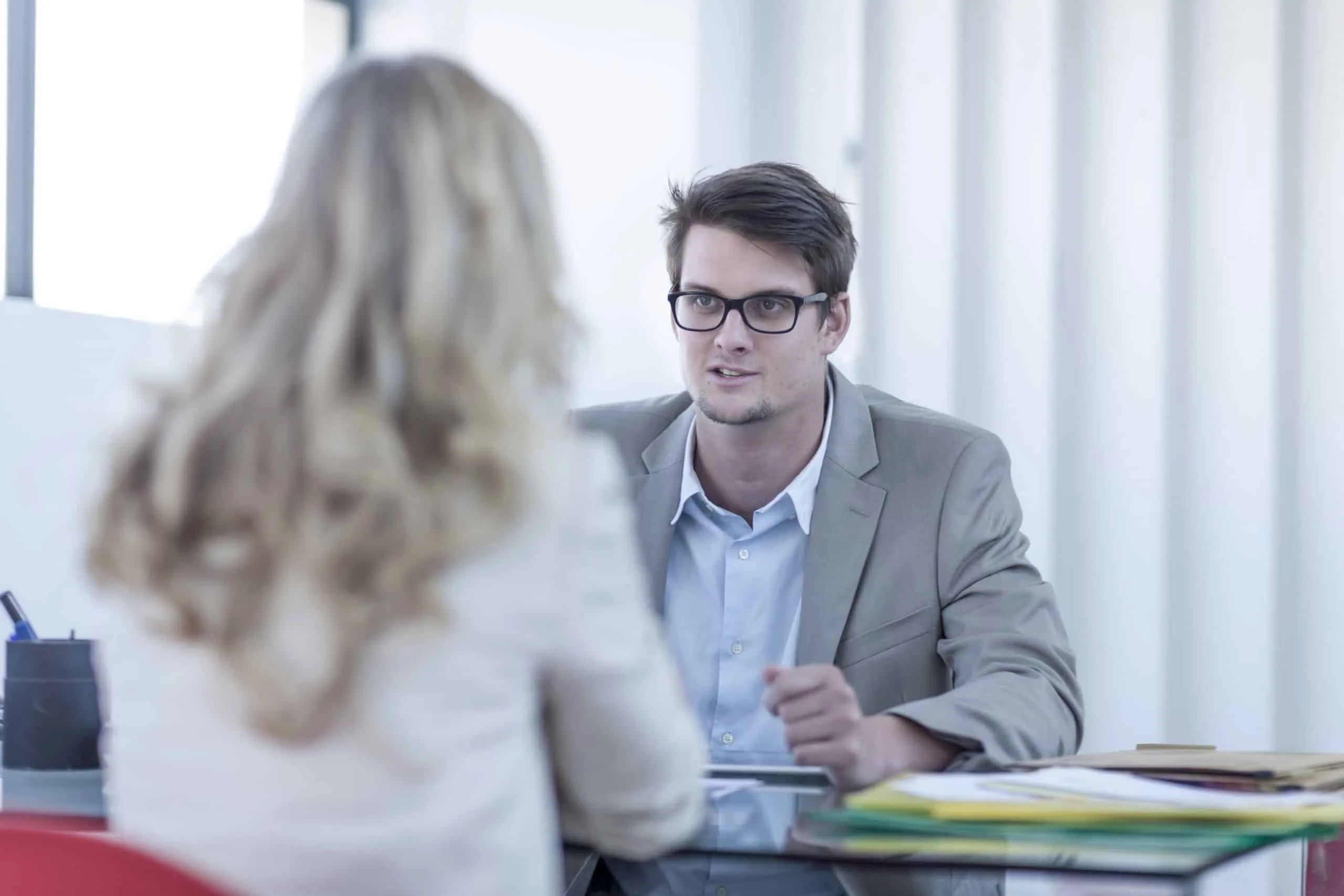Brünetter Mann mit Brille und hellem Anzug sitzt am Schreibtisch gegenüber einer blonden Frau, die mit dem Rücken zur Kamera zeigt und eine weiße Bluse trägt. Weißer verschwommener Hintergrund.