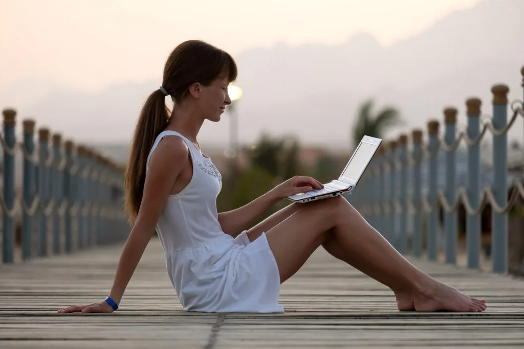 Brünette Frau im weißen Kleid hat Laptop auf dem Schoß und sitzt lächelnd an einem Steg. Verschwommener Hintergrund mit Dämmerung und Palmen.