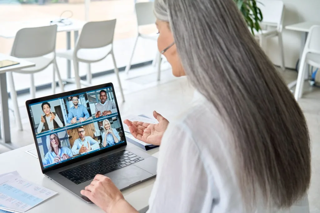 Grauhaarige Frau in weißer Bluse und Headset sitzt am Laptop und hält einen Video-Chat mit sechs Personen. Weiße Stühle und bodentiefe Fenster im Hintergrund.