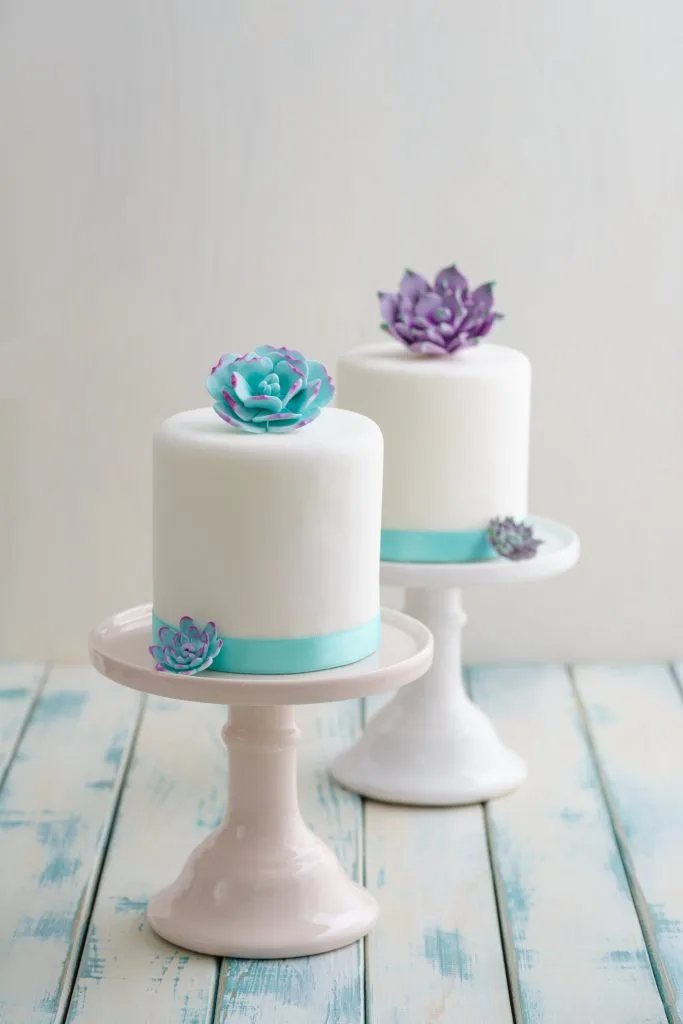 Zwei mit weißem Fondant und mit blauer sowie violetter Garnitur überzogene Mini-Torten auf weißen Tortenständern. Stehend auf blau-weißer Holzplattform.