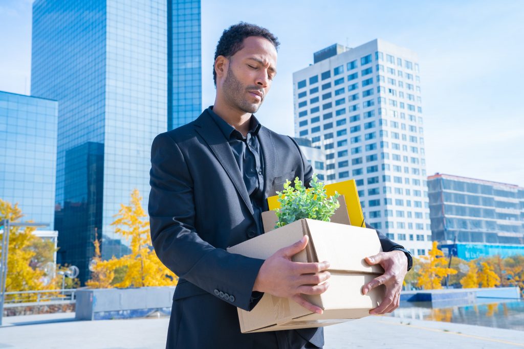 Enttäuscht dreinblickender schwarzhaariger Mann in dunkler Businesskleidung trägt braunen Pappkarton, in dem Ordner und eine Zimmerpflanze zu sehen sind. Im Hintergrund Hochhäuser, blauer Himmel und Bäume mit gelbem Herbstlaub.