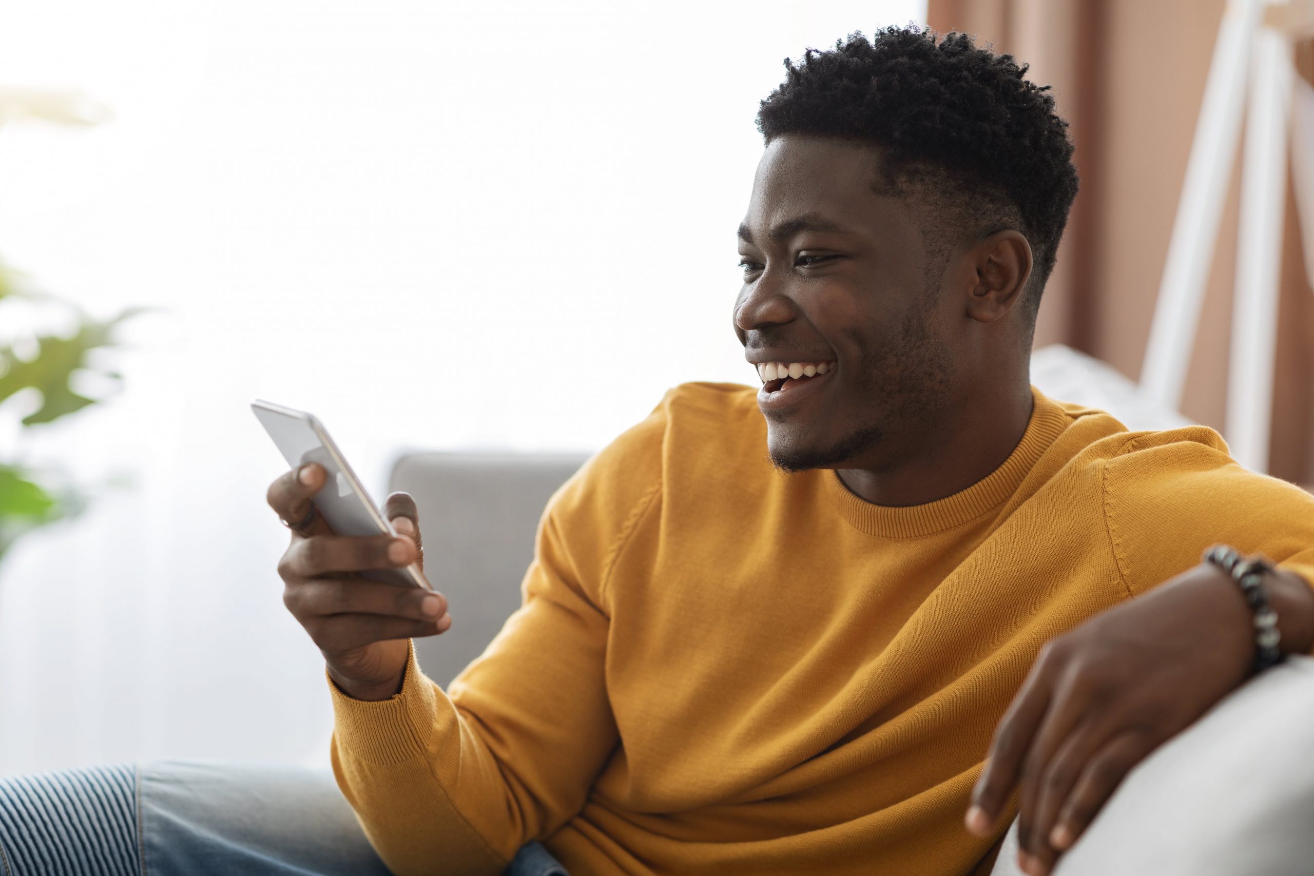 Lächelnder schwarzhaariger Mann in gelbem Pullover sitzt auf weißer Couch und guckt auf Handy.