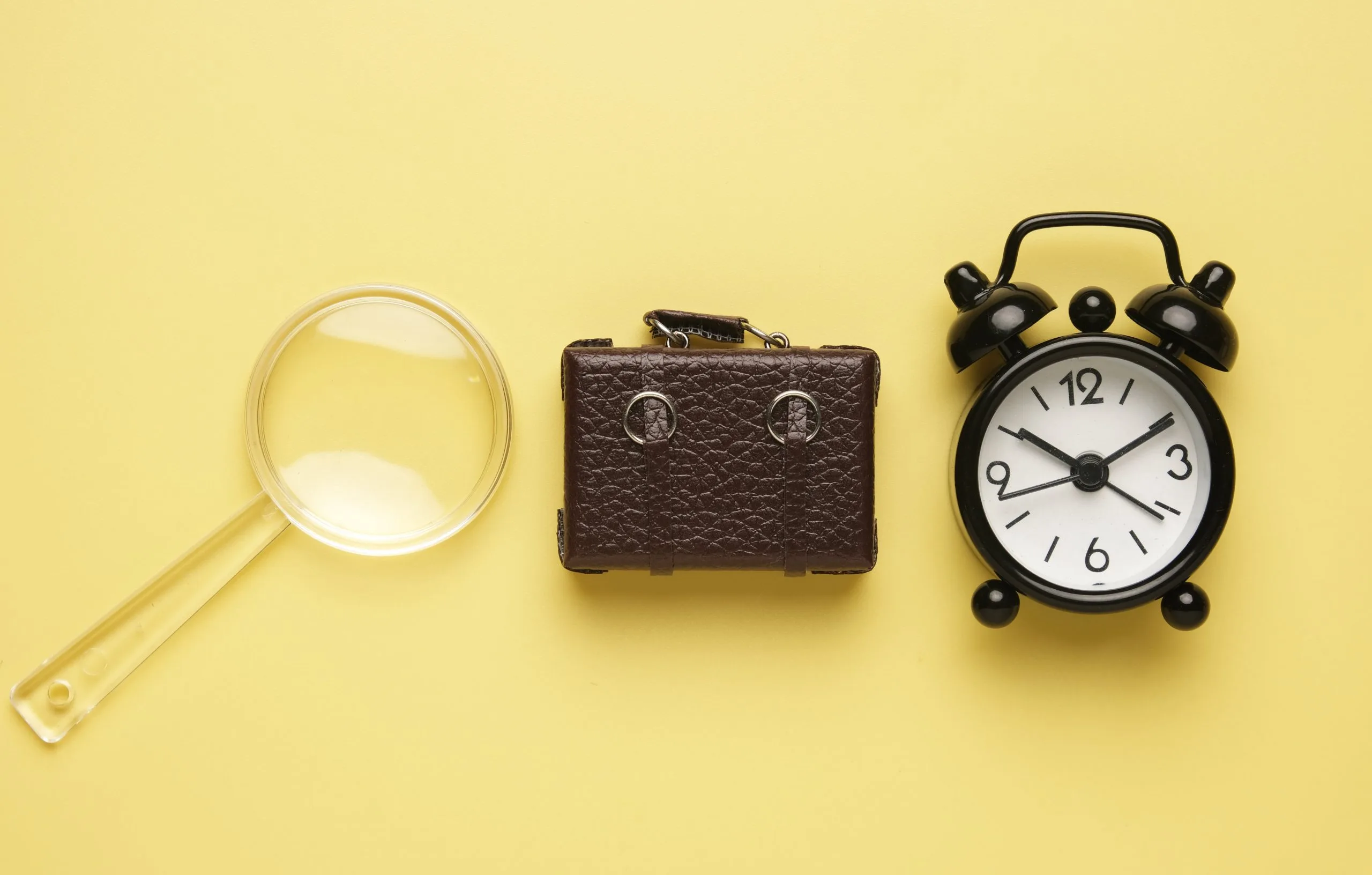 Miniatur einer Lupe, eines braunen Aktenkoffers und eines schwarzen Weckers vor gelbem Hintergrund.