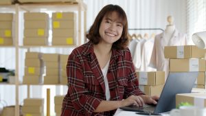Dunkelhaarige Frau in rotem kariertem offenem Hemd und weißem Top sitzt lächelnd am Laptop. Im Hintergrund Kartons in Regalen und Kleiderständer mit Kleidung.