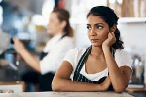 Frau mit dunklem Zopf, schwarz-weiß gestreifter Schürze und weißem T-Shirt steht an Verkaufstheke eines Cafés und hat das Kinn auf die Hand gestützt. Sieht unglücklich aus. Im Hintergrund verschwommen eine Kollegin, die Café verkauft.