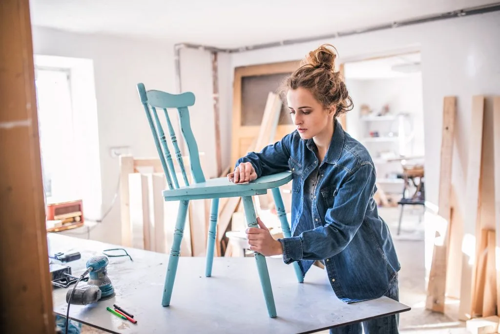 Blonde Frau mit Sleek-Dutt und Jeansjacke schreinert an einem blauen Stuhl. Schreinereizimmer mit Utensilien im Hintergrund.