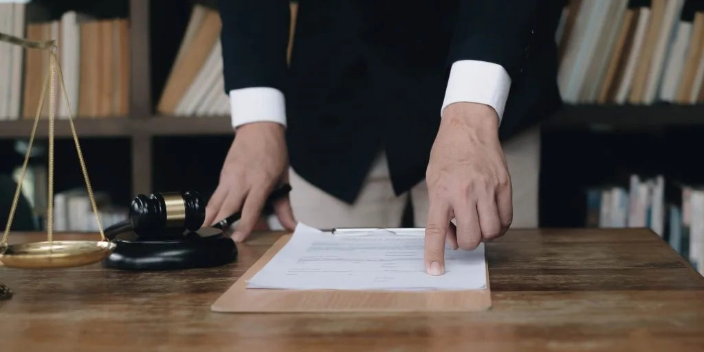 Mann, der von den Beinen bis zur Brust gezeigt wird, in dunklem Hemd und hellbrauner Hose, steht am Schreibtisch und hält einen Finger auf ein bedrucktes Blatt Papier. Daneben ein Richterhammer und ein Teil einer Waage. Bücherschrank im Hintergrund.