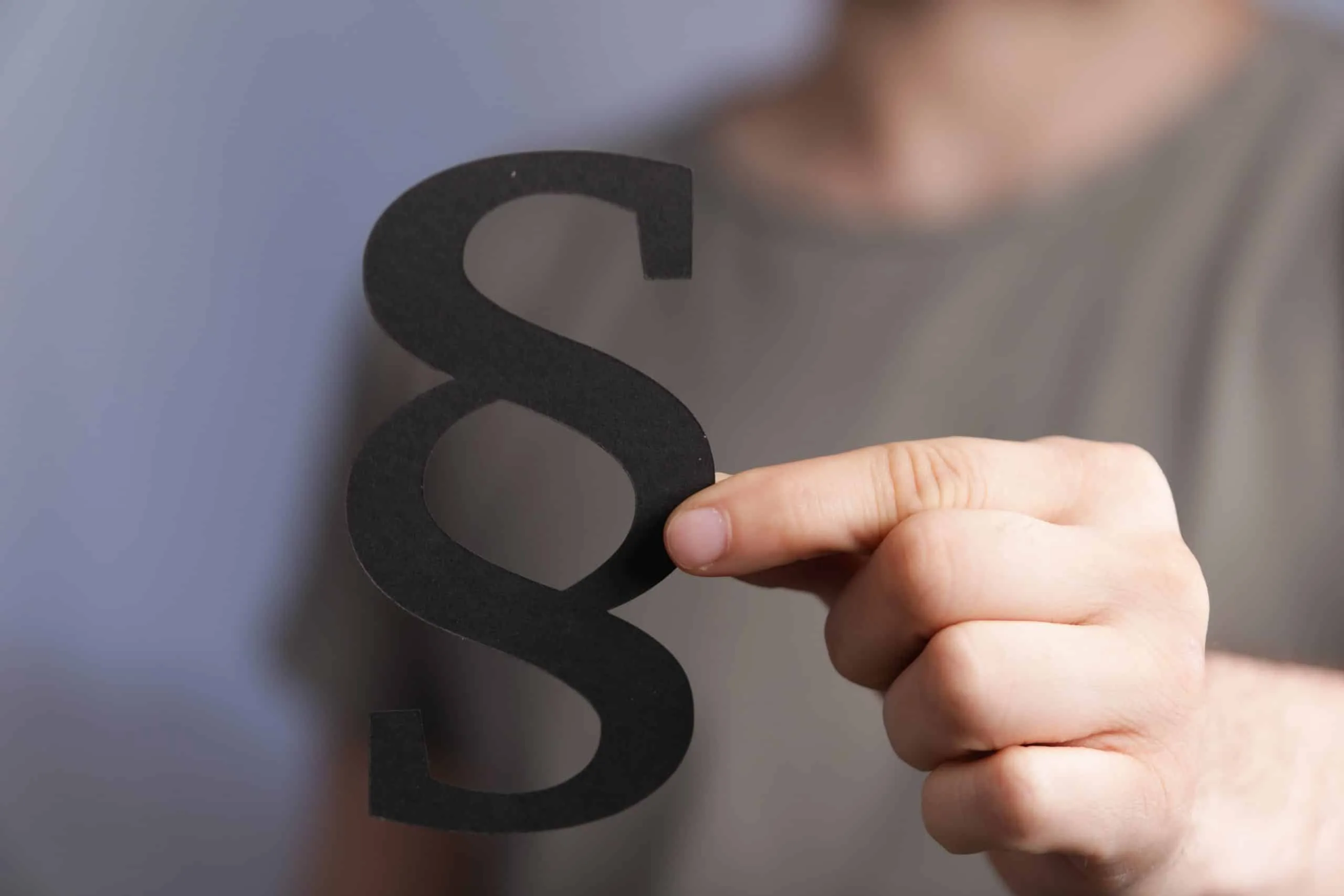 Männliche Hand hält ein schwarzes Paragraphenschild. Der verschwommene Brustbereich der Person im grauen T-Shirt im Hintergrund.
