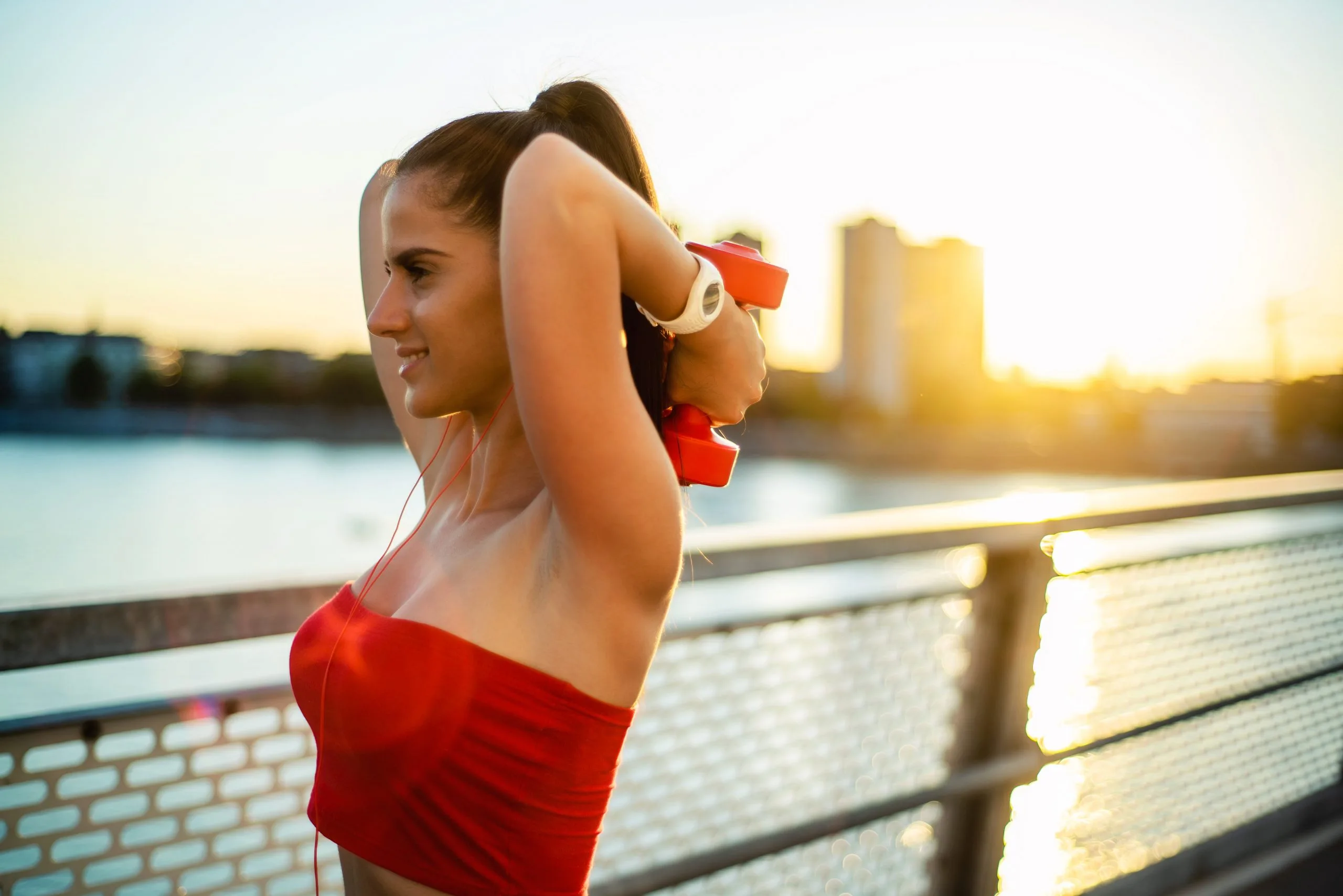 Brünette Frau mit Pferdeschwanz und rotem Top steht lächelnd mit roten Hanteln in den Händen an einem Brückengeländer und blickt auf einen Fluss hinaus. Im Hintergrund Dämmerung und Hochhäuser.