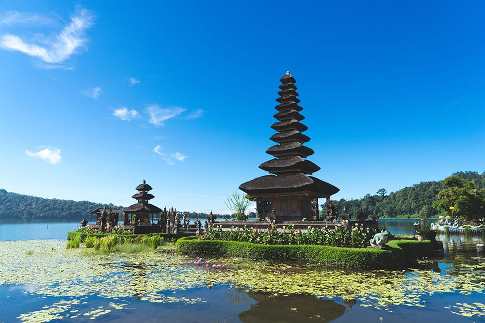 Schwimmender Tempel auf Bali bei blauem Himmel und Sonnenschein, umgeben von grünen Wasserpflanzen.