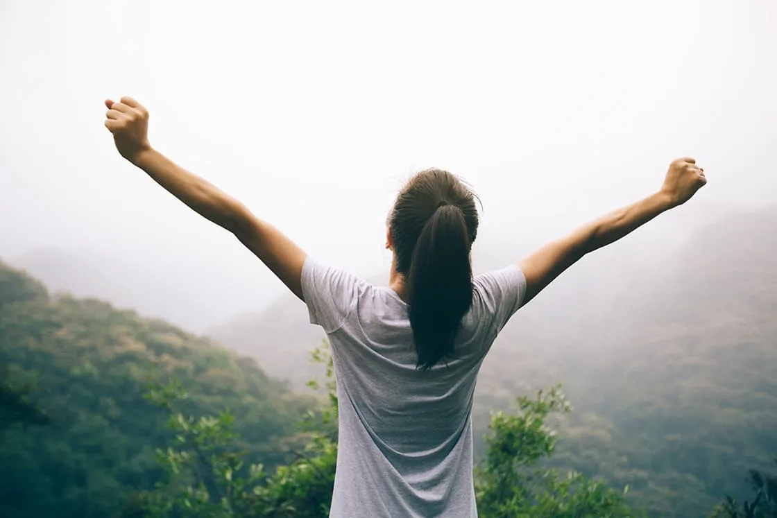 Brünette Frau mit Pferdeschwanz und weißem T-Shirt von hinten, hebt die Arme und steht vor einer Waldlandschaft.