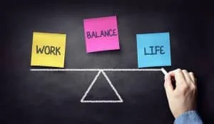 Drei Post-Its nebeneinander auf einer schwarzen Tafel– auf dem gelben steht Work, auf dem pinken steht Balance und auf dem Blauen steht Life geschreiben – darunter eine Hand, die mit Kreide eine Waage gezeichnet hat.