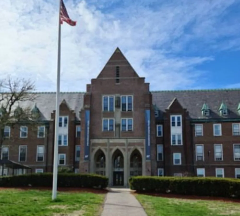 Älteres Gebäude mit Braunen Steinen, davor Gras, ein Steinweg, Gebüsch und USA-Flagge.