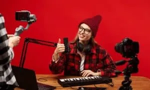 Blonde Bloggerin mit roter Mütze, rot-kariertem Hemd und Brille sitzt am Tisch mit Keyboard und Mikrophonen und hält den Daumen in Richtung Smartphone hoch, mit dem jemand sie filmt. Roter Hintergrund.