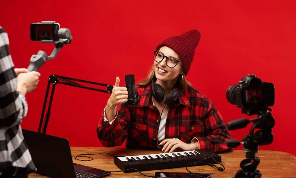 Blonde Bloggerin mit roter Mütze, rot-kariertem Hemd und Brille sitzt am Tisch mit Keyboard und Mikrophonen und hält den Daumen in Richtung Smartphone hoch, mit dem jemand sie filmt. Roter Hintergrund.