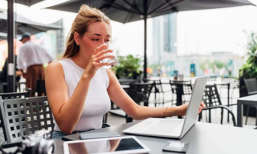 Blonde Frau in weißem Top sitzt im Außenbereich eines Restaurants mit Laptop und trinkt ein Glas Wasser.