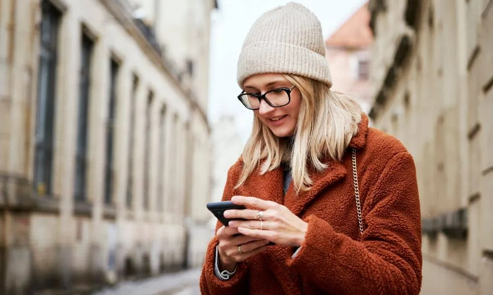 Blonde Frau mit Brille, weißer Mütze und brauner Plüschjacke ist in der Stadt am Smartphone.