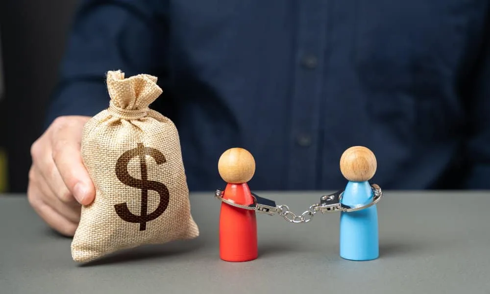 Brustbereich Mann in blauem Anzug, der einen Geldsack auf Schreibtisch hält. Daneben eine rote und eine blaue Holzspielfigur mit Handschellen.