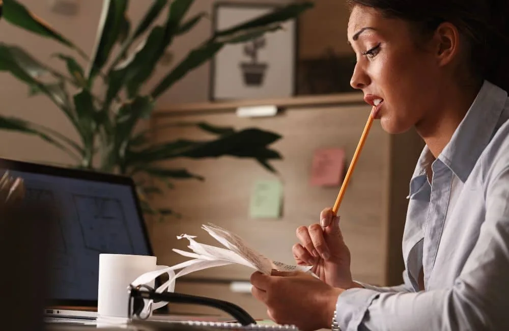 Dunkelhaarige Frau mit Bleistift im Mund am Schreibtisch grübelt über Dokumenten. Auf Schreibtisch Kaffeetasse, Zimmerpflanze im Hintergrund.
