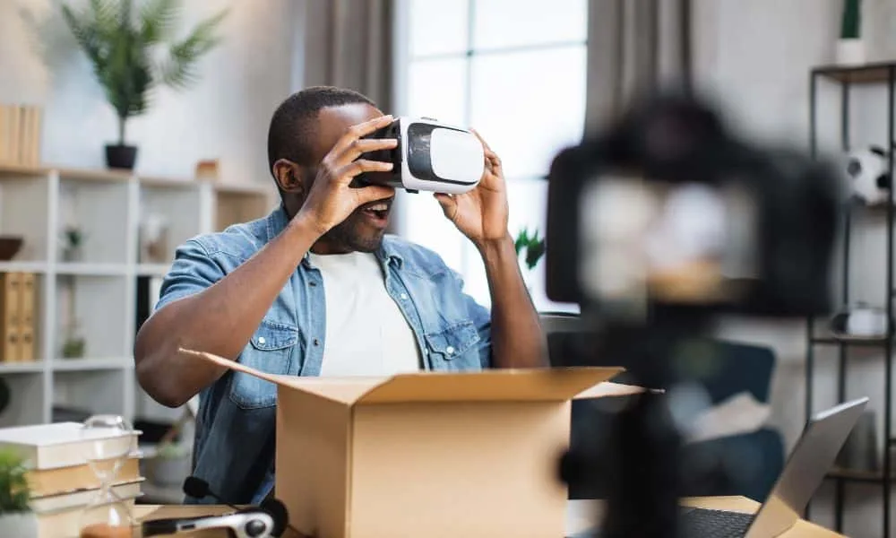 Dunkelhaariger Mann in Jeansjacke und weißem T-Shirt testet VR-Brille und nimmt sich dabei mittels Kameragestell auf. Weißes Regal mit verschiedenen Dingen im Hintergrund.