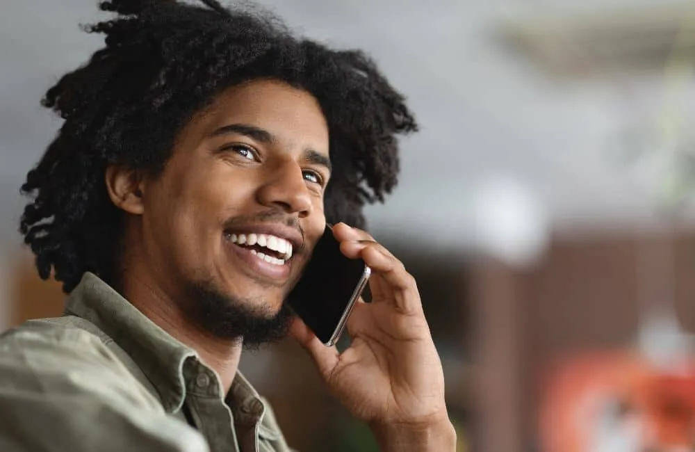 Dunkelhaariger Mann mit kurzem Kinnbart und in grauem Hemd bis zu dem Schultern lächelt während eines Handyanrufs. Stark verschwommener Hintergrund.