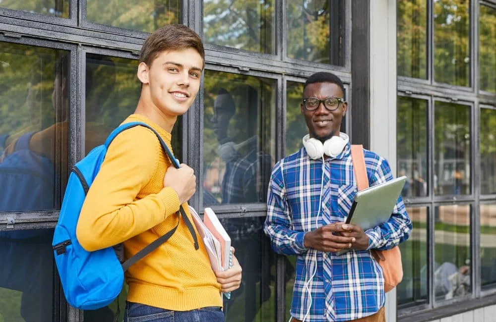 Ein dunkelblonder Mann in gelbem Pulli und mit blauem Rucksack steht bei einem schwarzhaarigen Mann in blau-weiß-kariertem Hemd mit Brille und weißen Kopfhörern um den Hals, der einen Laptop trägt. Im Hintergrund Gebäude mit zahlreichen Fenstern.